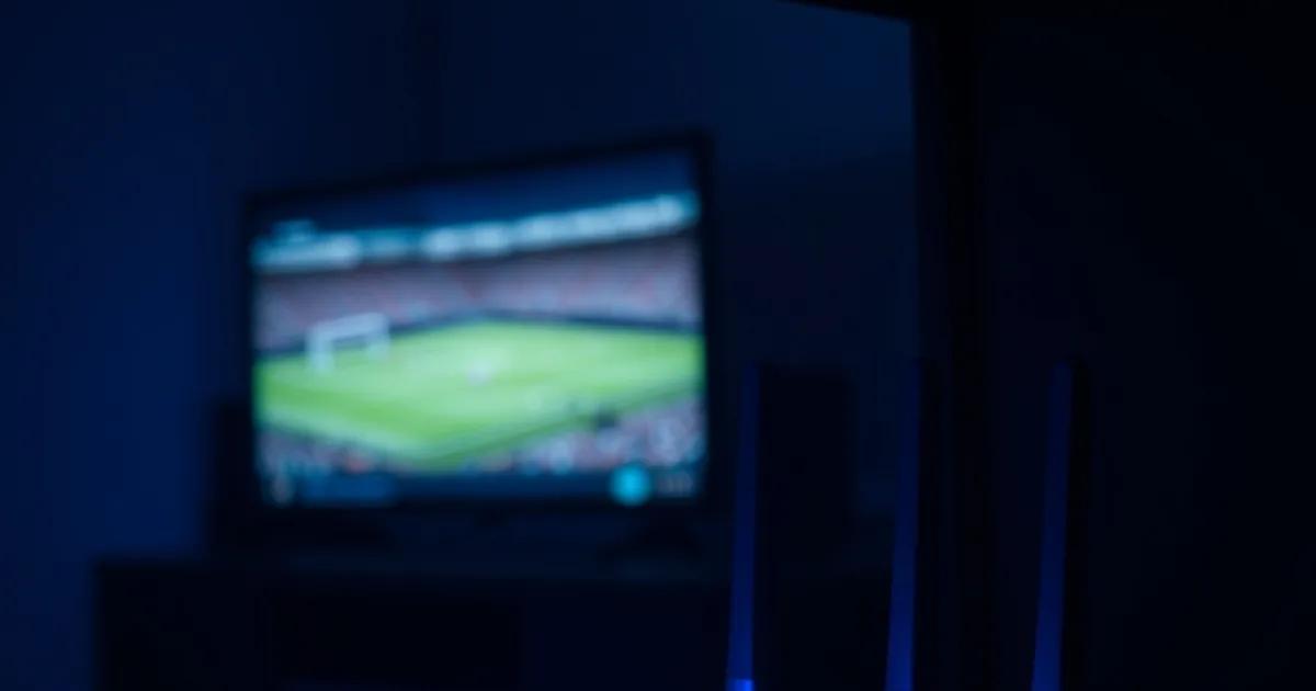 Close-up van een router met brandende statuslampjes, op de achtergrond een televisie die een voetbalwedstrijd uitzendt