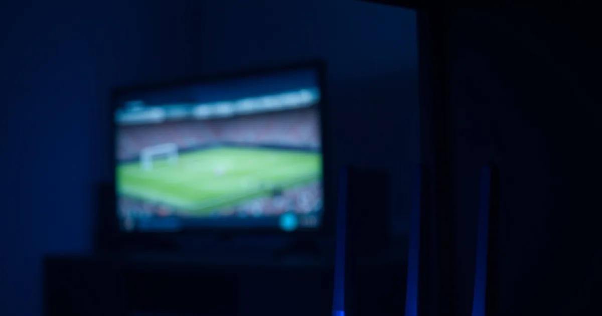 Close-up van een router met brandende statuslampjes, op de achtergrond een televisie die een voetbalwedstrijd uitzendt